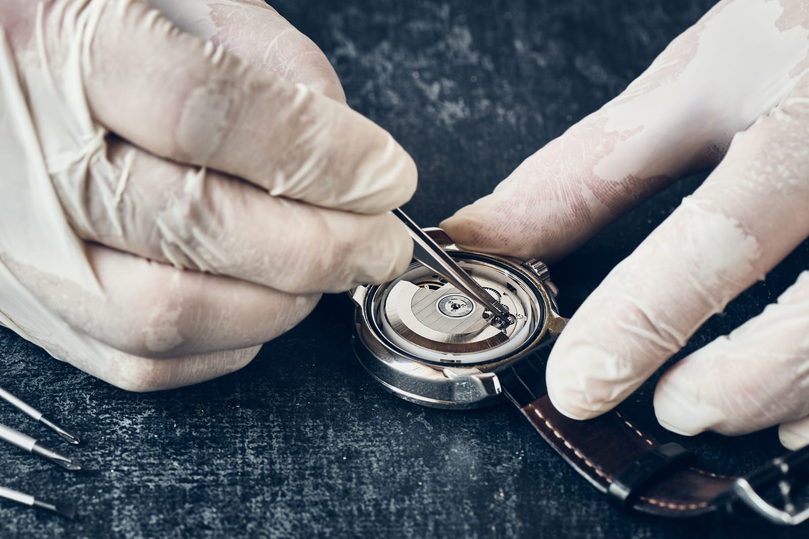 Mani con guanti che rimuovono il fondello di un orologio con carica a rotore, usando una pinzetta